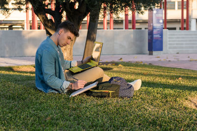 Smart concentrated man with backpack studying at laptop and writing on a notepad sitting in park grass with crossed legs in sunny day