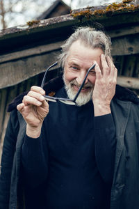 Elderly man with a beard stands in the village near a wooden shed in a sheepskin coat and glasses