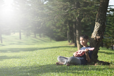 Boy playing guitar while sitting on grassy field at park