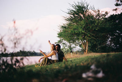 Portrait of smiling friends gesturing while sitting on grassy field by lake against sky