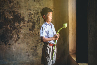 Boy holding flower bud while standing by window against wall