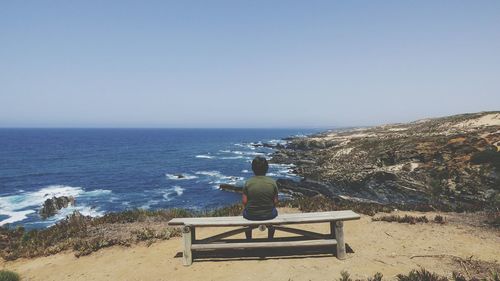 Rear view of boy relaxing on bench against sea