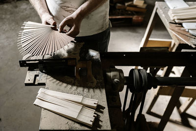 From above of unrecognizable crop craftsman working in dirty workshop at dusty workbench while standing with wooden folding fan