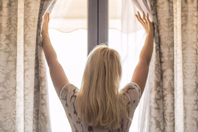 Rear view of young woman sliding window curtains at home
