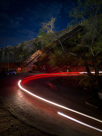 Light trails on road by trees against sky at night