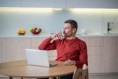 Freelancer man drinking water having break in work sitting at table on kitchen at home with laptop.