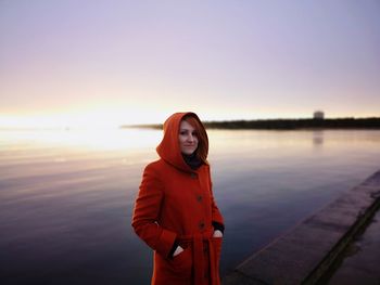 Portrait of woman wearing hooded shirt standing against sea during sunset