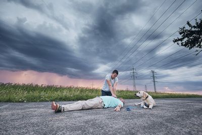 Man giving cpr to unconscious man by dog on road against cloudy sky