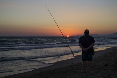 Rear view of man fishing on beach against sky during sunset
