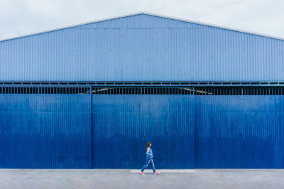 Remote view of female in denim apparel walking along street on background of blue building