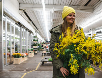 Woman in yellow cap and large bouquet of mimosa flowers at flower market
