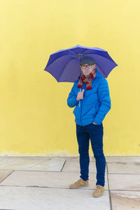 Confident senior man holding an umbrella standing against yellow wall