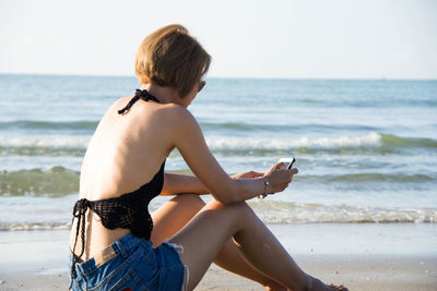 Side view of young woman using mobile phone while sitting at beach