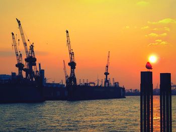 Silhouette cranes at harbor against sky during sunset