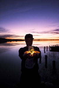 Man holding illuminated lights in glass jar while standing against sea and sky during sunset