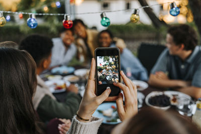 Woman photographing male and female friends through smart phone in back yard at dinner party