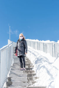 Adult woman with coat, sunglasses and a face mask standing on step of a staircase with a lot of snow