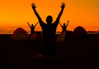 Silhouette people with arms raised at beach against orange sky