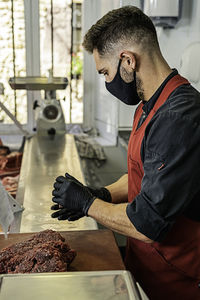 Butcher with mask making raw beef burgers in butchers shop.