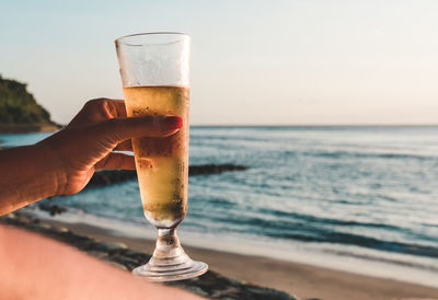 Hand holding glass of beer against sky at the beach 