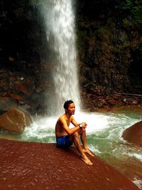 Young man sitting on rock against waterfall