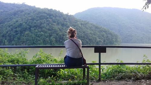 Rear view of woman sitting by river against tree mountains
