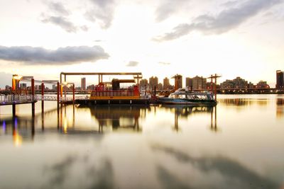 Pier on river by buildings against sky during sunset