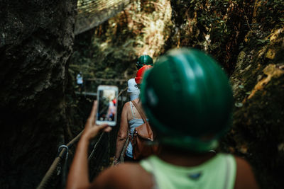 Back view of woman in safety helmet shooting on mobile phone tourists walking on footbridge between mountains