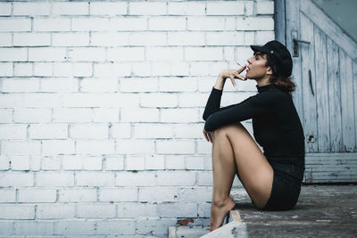 Young woman looking down while sitting against brick wall