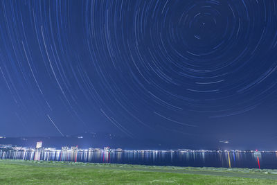 Scenic view of star trails over illuminated lake and grassy field