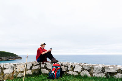 Full length side view of young female backpacker with trekking stick sitting on stone border near sea and browsing smartphone while resting and checking route during pilgrimage through spain