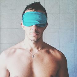 Shirtless young man wearing sleep mask while standing against tiled wall at home