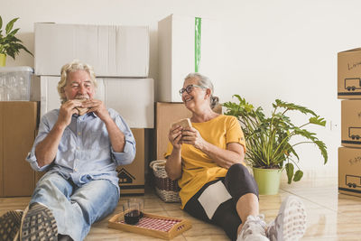 Full length of smiling senior couple eating while sitting on floor at home