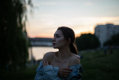 Young woman looking away while standing outdoors during sunset