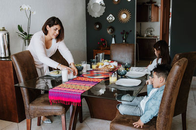 Side view of young latin american woman in casual clothes serving table with various food for children during breakfast