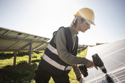 Female engineer using drill while working near solar panels at power station