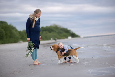 Pregnant woman standing by daughter playing with dog at beach against sky