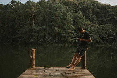 Man holding camera while sitting on bollard against lake