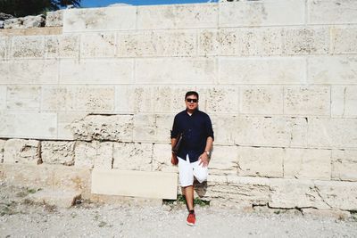 Full length portrait of smiling man standing against wall during sunny day