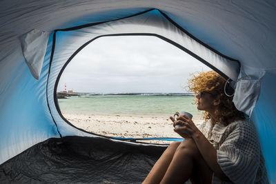 Woman drinking coffee while sitting in tent at beach