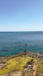 Mid distance view of man standing on rock by sea against blue sky
