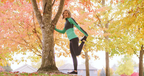 Young woman stretching leg while exercising by tree trunk in park during autumn