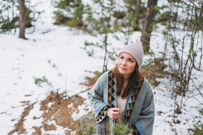 Portrait of young brunette beautiful woman in hat and grey poncho in winter forest