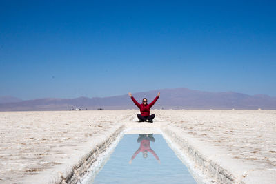 Woman with arms raised reflecting in water while sitting at salt flat