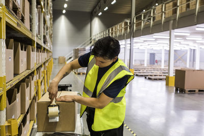 Young male worker packing cardboard box with adhesive tape at distribution warehouse