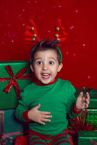 Baby boy in a green t shirt and with deer antlers sits with gifts on a red background