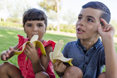 Two kids eating banana sitting outdoors on the grass in a park.