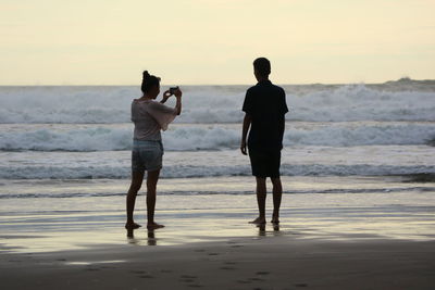 Rear view of friends standing on beach against sky during sunset