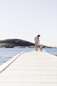 Rear view of mother standing with baby girl at the edge of wooden pier against clear sky