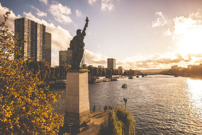 Statue of liberty replica by river against cloudy sky